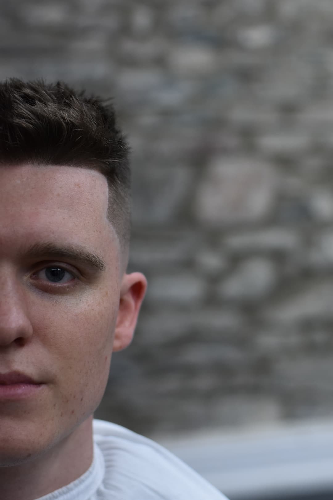 A freckled young man with short hair tilted sideways against a blurred stone background.